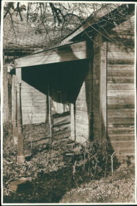 Black and white gelatin silver print of the front porch of an abandoned house with the wooden screen door standing open